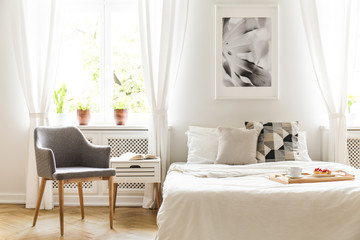 Grey armchair next to bed with wooden tray in bright bedroom interior with poster. Real photo