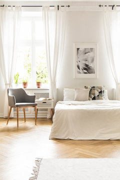 Grey armchair at window with drapes in white bedroom interior with poster above bed. Real photo