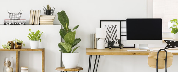Scandi home office interior with a desktop computer on a wooden desk next to a plant on a stool. Real photo with a mockup screen