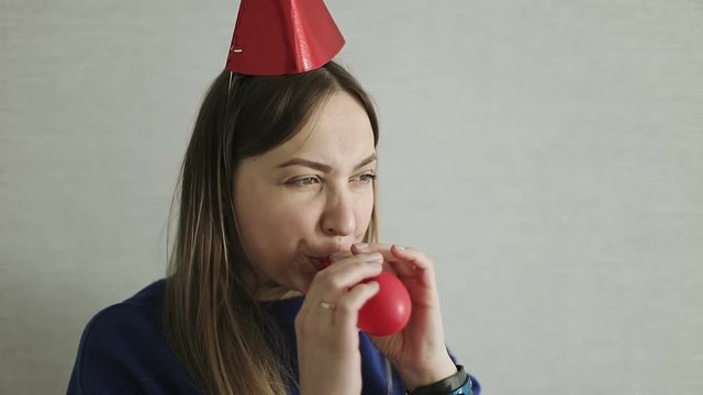 An adult woman in a festive hat inflates a red balloon indoors.