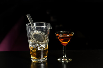 Closeup glass of Manhattan cocktail decorated with orange and decanter with ice at bar counter background.