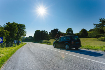 Cars driving on the asphalt road passing through green agricultural fields on a sunny day in Normandy, France. Countryside landscape, sunbeams in the blue sky, road network and transportation concept.