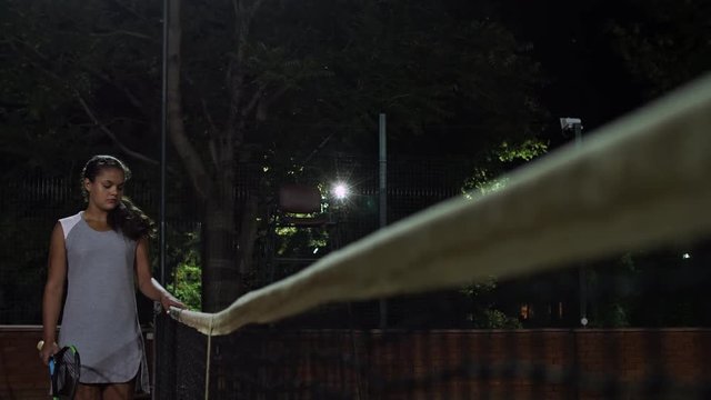 The girl tennis player walks along the tennis net. Playing tennis at night.
