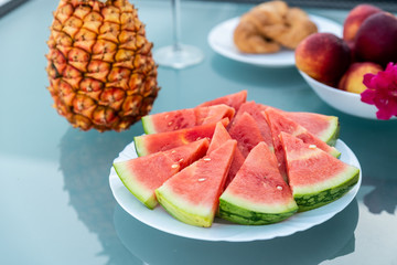 View of tasty breakfast on glass table, juice fruits, nectarine, watermelon, pineapple, water and bake. Healthy, colorful and exotic food in paradise. Summertime concept. Palm leaf on background.