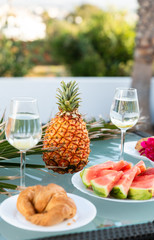 View of tasty breakfast on glass table, juice fruits, nectarine, watermelon, pineapple, water and bake. Healthy, colorful and exotic food in paradise. Summertime concept. Palm leaf on background.