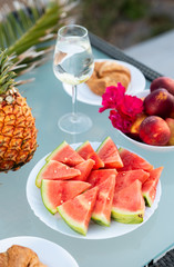 View of tasty breakfast on glass table, juice fruits, nectarine, watermelon, pineapple, water and bake. Healthy, colorful and exotic food in paradise. Summertime concept. Palm leaf on background.