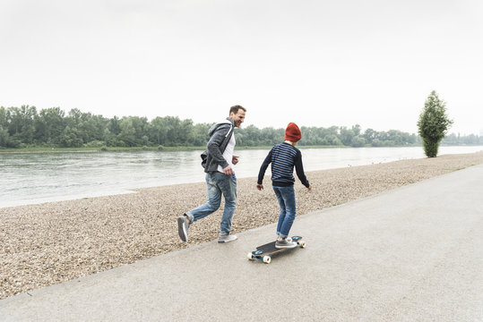 Happy father running next to son on skateboard at the riverside