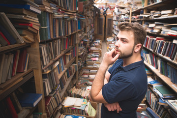 Portrait of a man with a beard standing on the background of an atmospheric public library and looking at the bookshelf.Student selects books in a library.Searching for literature in a public library