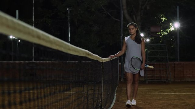 The girl tennis player walks along the tennis net. Playing tennis at night.