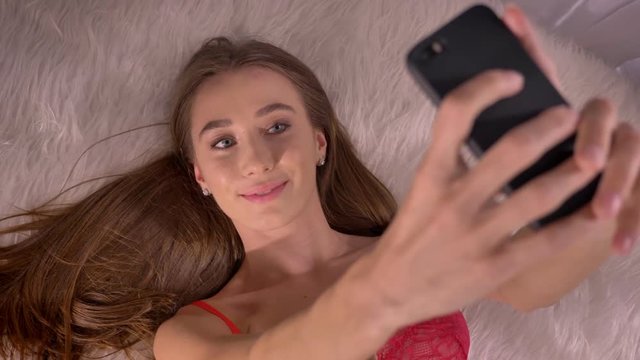 Young attractive woman in red bra lying on bed with fur and taking selfie, sexy model with blue eyes taking photos of herself