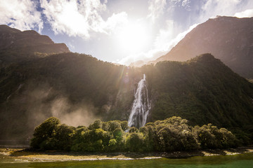 a beautiful natural landscape of green mountains with waterfall falling from mountain to small forest on ground  in the lake with sunlight, fog and sunbeams