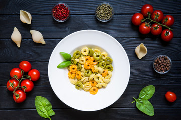 Assortment of tortellini, red tomato, green with spinach and cheese. Black wooden background with cherry tomatoes, allspice, red and green pepper in glass bowls. italian cuisine on rustic background.
