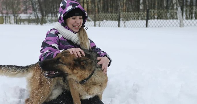 A young woman in a winter jacket plays with a German shepherd in the snow.
