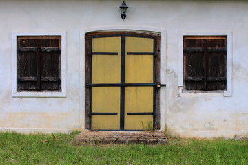 Obraz na płótnie Canvas Renovated massive wooden doors locked with padlock surrounded with two closed wooden blinds on small windows and old lamp on top with stone entrance and uncut grass in front