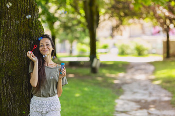 Gorgeous young brunette girl blowing soap bubbles in sunlit park.