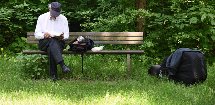 Auf einer Parkbank im Gr&uuml;nen sitzender Cellist in wei&szlig;em Hemd mit Fliege und einem Buch in der Hand