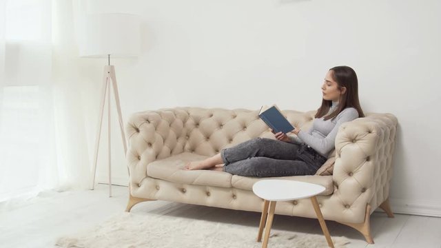 Young caucasian female reading book on sofa in white room with floor lamp and window.