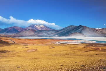 Naklejka premium Pasmo górskie Piedras Rojas w pobliżu San Pedro de Atacama, Chile. Przed kolorową górą skalistą znajduje się półsuche słone jezioro lub salar o intensywnym turkusowym kolorze. Przy autostradzie jechał czerwony samochód.