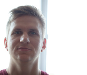 A young man seriously looks at the camera standing near the window in the room