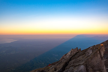 A Man In Red Jacket is Climbing to The Top of Mount Agung Volcano, The Active Volcano in Bali, Indonesia at Sunrise with A Shadow of Mount Agung Over Bali Background.