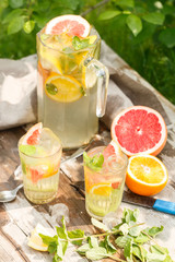 citrus lemonade in glass jug with mint. Orange. Grapefruit and lemon. On greenery background.