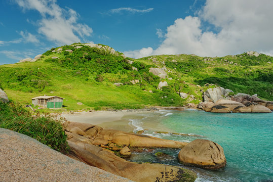 Pequena praia deserta com casebre de madeira, colinas verdes, rochas e mar azul turquesa