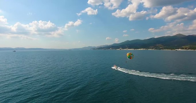 Aerial flight speedboat floats on surface of azure sea and pulls parasail with tourist through air on pictorial background with mountains and coast strip under blue sky with white clouds. Parasailing