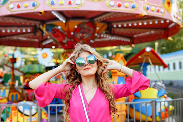 Obraz na płótnie Canvas Bright summer concept. A cheerful blonde girl in pink jacket and stylish sunglasses is having fun in the amusement Park. Cheerful woman smiling against bright carousel