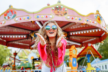 blonde girl in pink jacket and stylish sunglasses is having fun in the amusement Park. The woman smiles cheerfully and pulls her hands to us against the background of a bright carousel