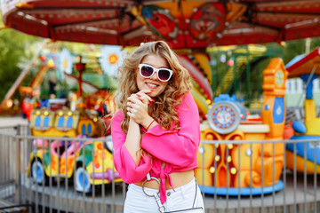 Bright summer concept. A cheerful blonde girl in pink jacket and stylish sunglasses is having fun in the amusement Park. Cheerful woman smiling against bright carousel