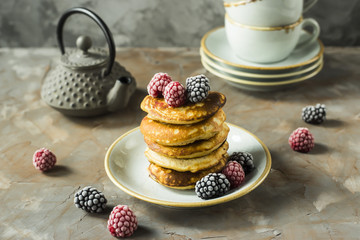 A pile of pancakes on a plate with raspberries and blackberries next to cups and a kettle on a gray table. Summer Breakfast concept