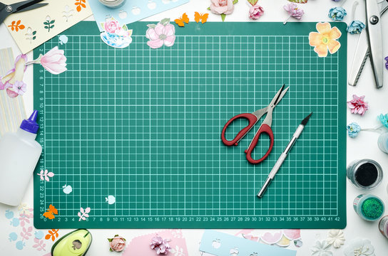 The cutting mat is surrounded by paper flowers, paper, tools and scrapbooking materials. Scrapbooking, top view