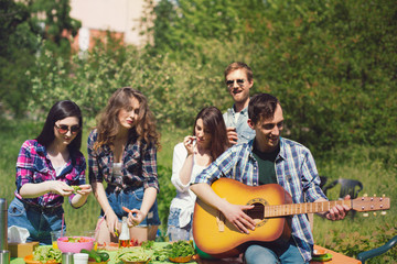 Company of friends enjoying time together on picnic. Guy playing guitar sitting on table out on picnic with his friends in background.