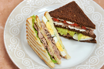On the plate are two triangular-shaped sandwiches. One sandwich of white bread, the other of dark. Filling of tuna sandwiches, hard boiled eggs and vegetables. Light background. Close-up.