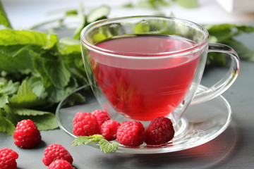 Transparent Cup of fragrant tea with addition of berries of a raspberry on a dark background, the scattered around twigs of raspberry and mint leaves