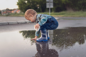 Toddler boy playing in puddle outdoors after rain wearing rubber boots, crouching and touching water. Outdoors, natural lighting, no retouch, matte filter.