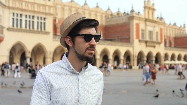 front view of stylish traveling man in hat sunglasses walking and looking around on city square with pedestrians people on blur background summer vacation sightseeing enjoying beautiful architecture