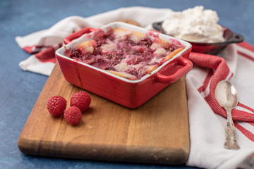Homemade Raspberry Cobbler in Red Cookware on Wooden Cutting Board beside a Red & White Kitchen Towel on a Blue Background