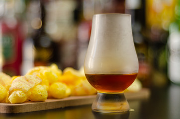 glass of whiskey with ice cubes and salty snacks on the background of bottles and bar