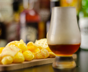 glass of whiskey with ice cubes and salty snacks on the background of bottles and bar