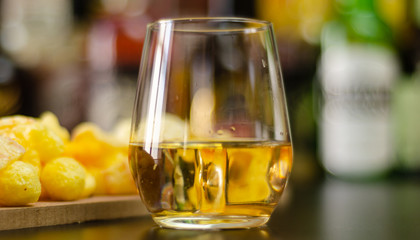 glass of whiskey with ice cubes and salty snacks on the background of bottles and bar