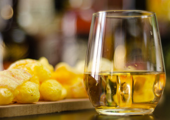 glass of whiskey with ice cubes and salty snacks on the background of bottles and bar