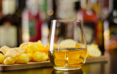 glass of whiskey with ice cubes and salty snacks on the background of bottles and bar