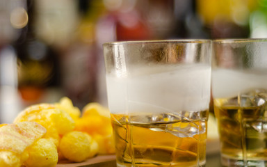 glass of whiskey with ice cubes and salty snacks on the background of bottles and bar