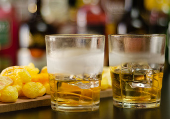 glass of whiskey with ice cubes and salty snacks on the background of bottles and bar