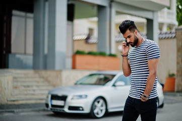 Fototapeta na wymiar Handsome tall arabian beard man model at stripped shirt posed outdoor against car. Fashionable arab guy. Muslim businessman speaking at mobile phone.