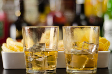 glass of whiskey with ice cubes and salty snacks on the background of bottles and bar