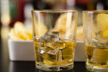 glass of whiskey with ice cubes and salty snacks on the background of bottles and bar