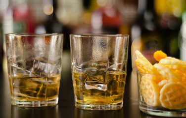 glass of whiskey with ice cubes and salty snacks on the background of bottles and bar