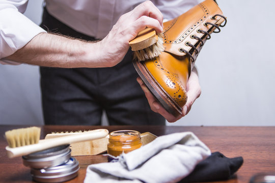 Hands of Professional Male Footwear Cleaner Using Brush For Polishing Male Tan Brogue Derby Boots. Variety of Cleaning Accessories Used.